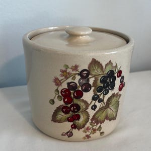 May include: A ceramic jar with a lid, featuring a floral design with red and black berries. The jar is cream-colored with a textured surface.