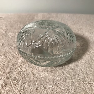 May include: A clear glass trinket box with a decorative, geometric pattern. The box is round and has a lid.