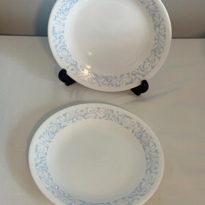 May include: Two white ceramic plates with a light blue floral pattern border. The plates are round and have a simple, elegant design. One plate is on a black stand. The plates are clean and appear to be in good condition.