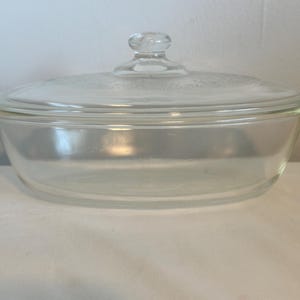 May include: Clear glass oval baking dish with a matching lid. The dish has a simple design with a slightly textured lid and a small knob handle. The dish is designed for oven use.
