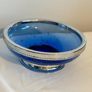 May include: A blue glass bowl with a silver rim and base. The bowl has a rounded shape and a deep blue color. The silver rim and base add a touch of elegance to the design. The bowl is likely used for serving or display.
