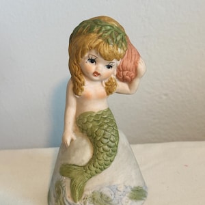 May include: A ceramic mermaid figurine with blonde hair and a green tail. She holds a pink seashell to her ear. The figurine is painted with delicate details and sits on a base with painted waves.