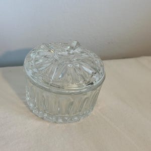 May include: A clear, round glass jar with a lid. The jar has a textured, ribbed design on the sides and a decorative, starburst pattern on the lid. The lid has a small handle. The jar is sitting on a white surface.