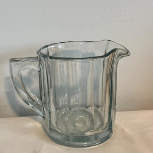 May include: A clear glass pitcher with a handle and a pouring spout. The pitcher has vertical ribbed detailing and a decorative circular pattern on the base. The pitcher is designed for serving beverages.
