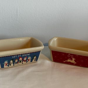 May include: Two ceramic loaf pans with festive holiday designs. One pan features a blue and white snowman design with the text "Let It Snow". The other pan has a red and white reindeer design.
