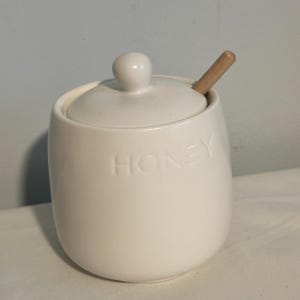 May include: A white ceramic honey pot with a lid and a small wooden honey dipper. The word "HONEY" is embossed on the front of the pot. The pot is sitting on a white surface.