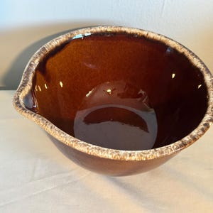 May include: A brown ceramic mixing bowl with a glossy finish. The bowl has a unique shape with a spout and a textured, light-colored rim. This vintage kitchenware is ideal for mixing ingredients.