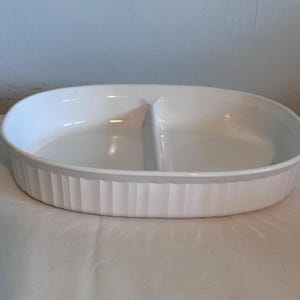 May include: A white oval ceramic serving dish featuring a central divider that splits the interior into two equal sections. The exterior of the dish is decorated with a vertical fluted or ribbed pattern around the entire perimeter of the bowl.