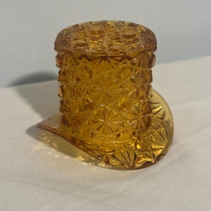 May include: A small, amber-colored glass top hat with a textured, starburst pattern. The hat has a cylindrical crown and a wide, flat brim. The glass is transparent, allowing light to pass through, and is likely a decorative item.
