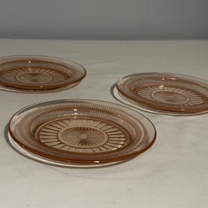 May include: Three pink glass dessert plates with a raised, circular design in the center. The plates are stacked on a white surface.