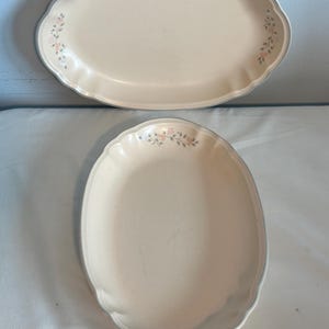 May include: Two oval serving platters. The platters are cream-colored with a scalloped edge and a light blue trim. Floral accents in pink and green are on each end of the platters. The platters are made of a durable material.