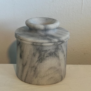 May include: A cylindrical marble butter dish with a lid and a small handle. The dish is gray with darker gray veining throughout. The lid fits snugly on top of the dish, creating a cohesive and functional design.