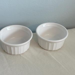 May include: Two white, round ramekins with vertical ridged sides. The ramekins are empty and sit side-by-side on a white surface. The background is a light blue wall.