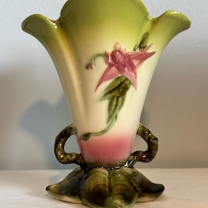 May include: A vintage ceramic vase with a flared top and a floral design. The vase features a light green rim, a cream-colored body with a pink base, and a painted pink flower with green leaves. It has two brown handles and a leaf-shaped base.