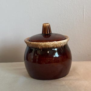 May include: A small, round, brown ceramic jar with a lid. The jar has a glossy finish and a light brown rim. The lid has a small, ridged handle. The jar is likely used for storing food items.