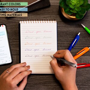 May include: A hand writing "Draw your dreams" on a lined notebook with a silver pen. There are four colorful pens on a wooden table next to the notebook.