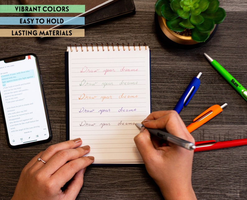 May include: A hand writing "Draw your dreams" on a lined notebook with a silver pen. There are four colorful pens on a wooden table next to the notebook.
