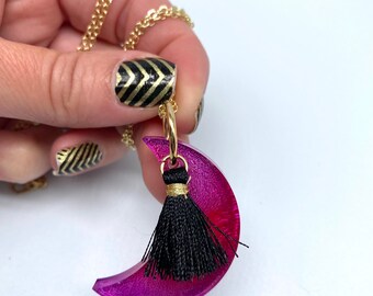 May include: A pink crescent moon pendant necklace with a black tassel. The necklace is made of resin and has a gold chain.