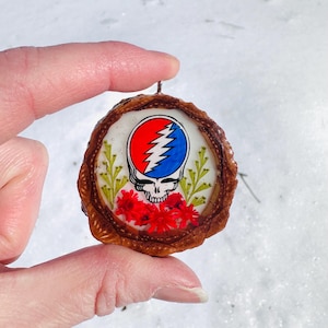 May include: A round pendant featuring the Grateful Dead's Steal Your Face logo in red, white, and blue, surrounded by a brown wooden frame. The pendant includes red flowers and green foliage. The pendant is held up by a person's fingers.