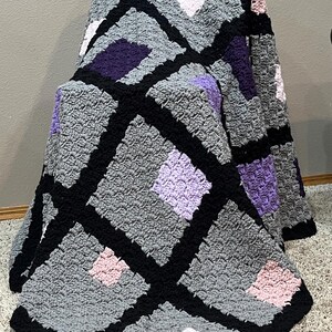 May include: A crocheted blanket with a diamond pattern in shades of gray, purple, pink, and white. The blanket is bordered in black.