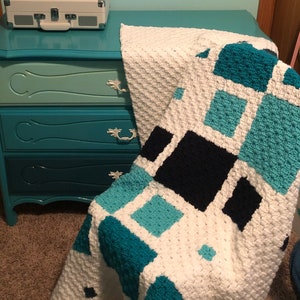 May include: A white crocheted blanket with blue and black squares. The blanket is draped over a teal dresser with a white vintage record player on top.