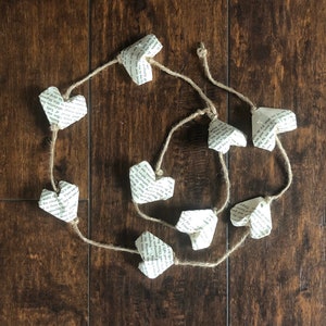 Puede incluir: Una guirnalda de corazones de papel hechos con páginas de libros, atados con un cordel. Los corazones son de diferentes tamaños y formas, y son de color crema claro.