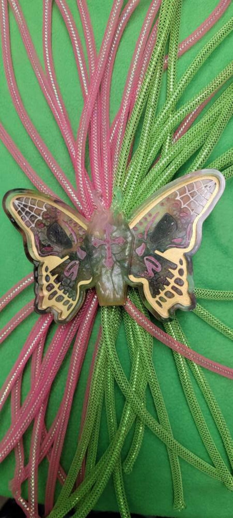 May include: A green and pink butterfly-shaped resin charm with a detailed design. The charm is surrounded by pink and green sparkly cords.