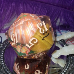 May include: A large, multi-faceted die with a pumpkin and autumn leaf design inside. The die has white numbers and a yellow, smiling face on one side. The die is reflected in a mirror with a decorative frame.