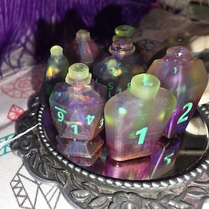 May include: A collection of translucent, multi-faceted resin potion bottle dice with iridescent purple and pink internal swirls. Each bottle-shaped die features bright neon green numbers and a small, cylindrical stopper top, arranged on a silver mirror.