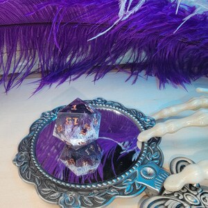 May include: A purple and white iridescent dice with gold numbers sits in a silver ornate hand mirror with a purple background. The mirror is on a white surface.