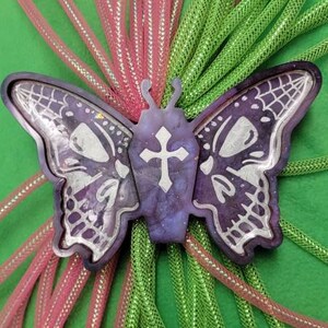 May include: A purple and silver butterfly with a cross on its chest, surrounded by pink and green mesh fabric.