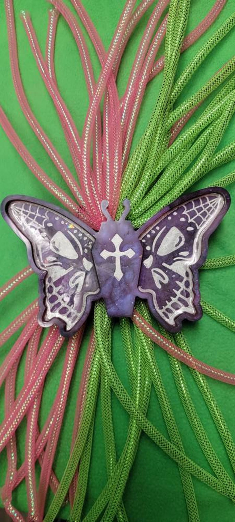 May include: A purple and silver butterfly with a cross on its chest, surrounded by pink and green mesh fabric.
