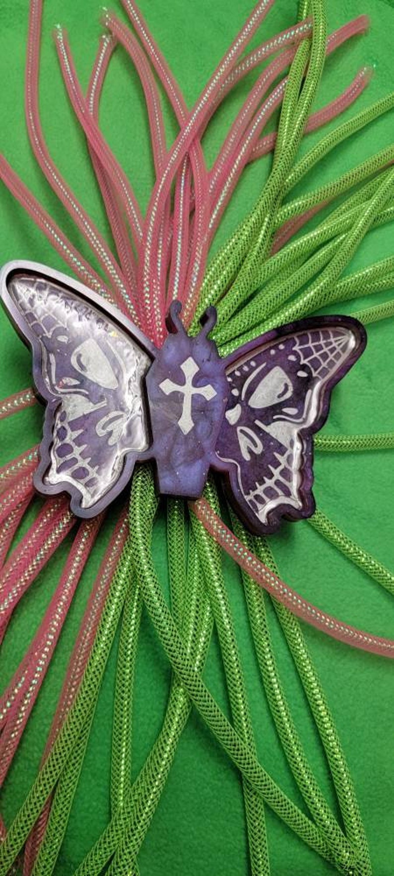 May include: A purple resin butterfly with a cross design, surrounded by pink and green sparkly mesh ribbon.