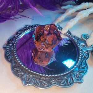 May include: A purple and gold resin dice with a 20 on one side, sitting in a silver ornate mirror with a purple feather in the background.
