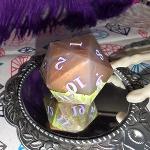 May include: A twenty-sided die featuring a translucent brown resin body with embedded gold glitter and light green accents at the base. The die has engraved numbers painted in a light purple color, resting on a small, ornate, circular silver mirror.