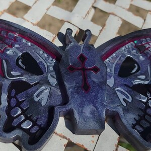 May include: A purple and silver resin butterfly with a black cross on the body and a skull design on the wings. The butterfly is on a white surface.
