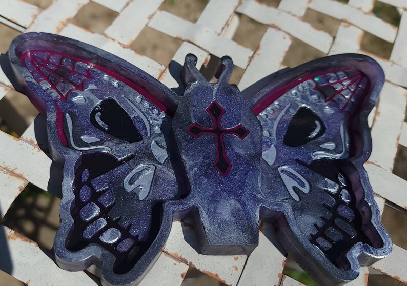May include: A purple and silver resin butterfly with a black cross on the body and a skull design on the wings. The butterfly is on a white surface.