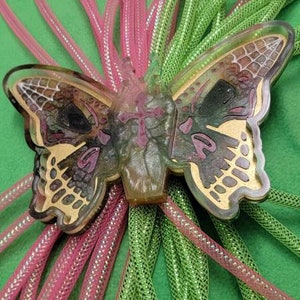 May include: A green and pink iridescent butterfly charm with a glittery finish. The charm is attached to a bundle of green and pink iridescent cording.