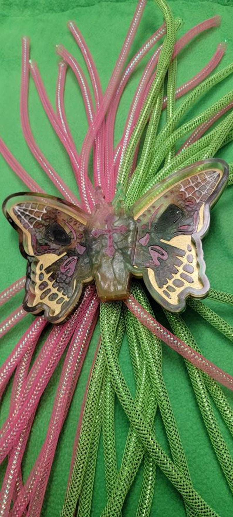 May include: A green and pink iridescent butterfly charm with a glittery finish. The charm is attached to a bundle of green and pink iridescent cording.