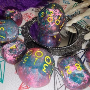 May include: A collection of handmade, galaxy-themed dice with yellow numbering. The dice are a mix of pink, blue, and purple with glitter accents. Some dice are reflected in a small, ornate mirror.