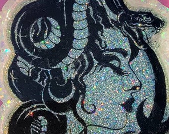 May include: A black and white resin art piece featuring a woman's face with a snake wrapped around her head. The background is a glittery silver color.