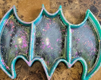 May include: A teal blue resin tray shaped like a bat with three compartments. The tray is filled with black and iridescent glitter.