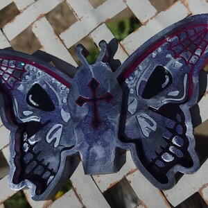 May include: A silver and black butterfly-shaped resin tray with a cross design in the center. The butterfly wings have a skull design with a red border.