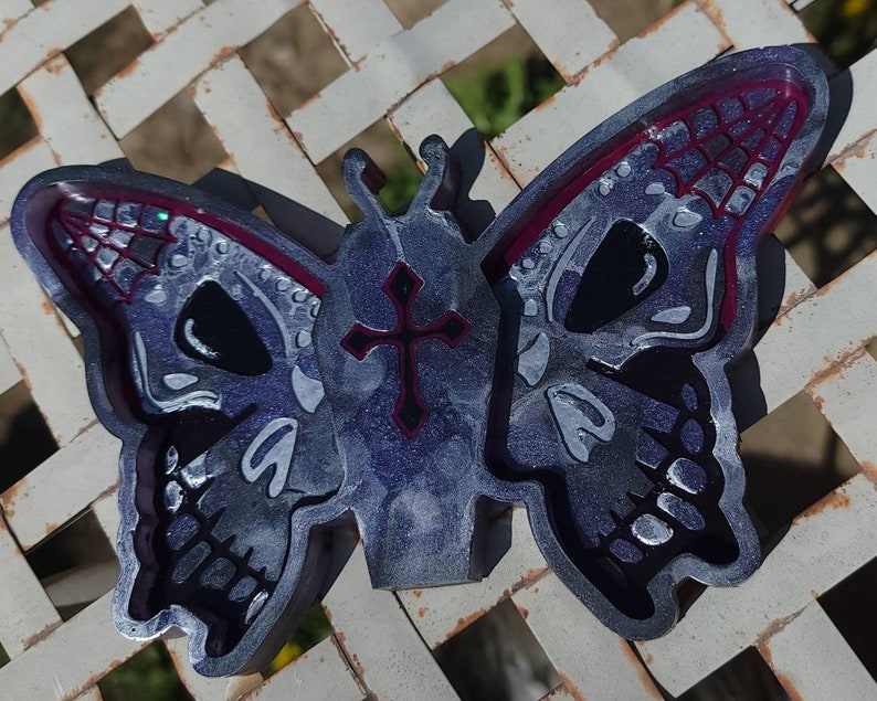 May include: A silver and black butterfly-shaped resin tray with a cross design in the center. The butterfly wings have a skull design with a red border.