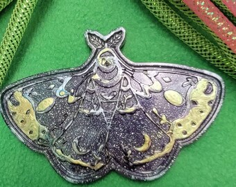 May include: A black and gold glitter resin moth pin with intricate details and a celestial design. The moth has a dark purple body with gold accents and a pair of wings with a crescent moon design.