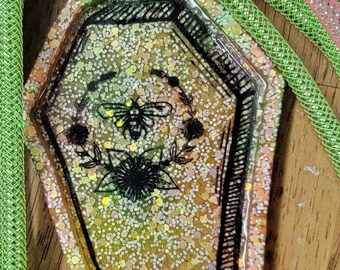 May include: A small, coffin-shaped trinket dish with a black outline and a glittery, iridescent finish. The dish features a black and white illustration of a bee and flowers.