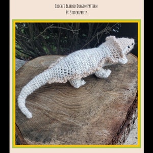 May include: A crocheted bearded dragon toy in beige and white, sitting on a wooden surface. The toy is posed with its head turned to the side and its tail curled behind it.