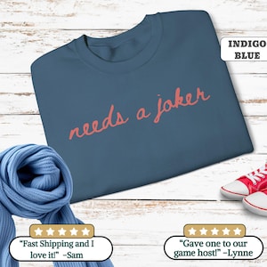 May include: Indigo blue sweatshirt with the text "needs a joker" in coral lettering. The sweatshirt is displayed on a wooden surface, with a blue scarf and red sneakers in the foreground. The text "INDIGO BLUE" is in the upper right corner.