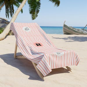 May include: A beach towel with a coral and white vertical striped pattern draped over a wooden lounge chair. The towel features a circular emblem with a black letter R in the center, flanked by small black line art icons of a book and a camera.