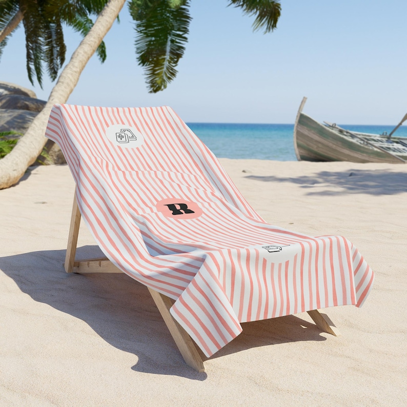 May include: A beach towel with a coral and white vertical striped pattern draped over a wooden lounge chair. The towel features a circular emblem with a black letter R in the center, flanked by small black line art icons of a book and a camera.